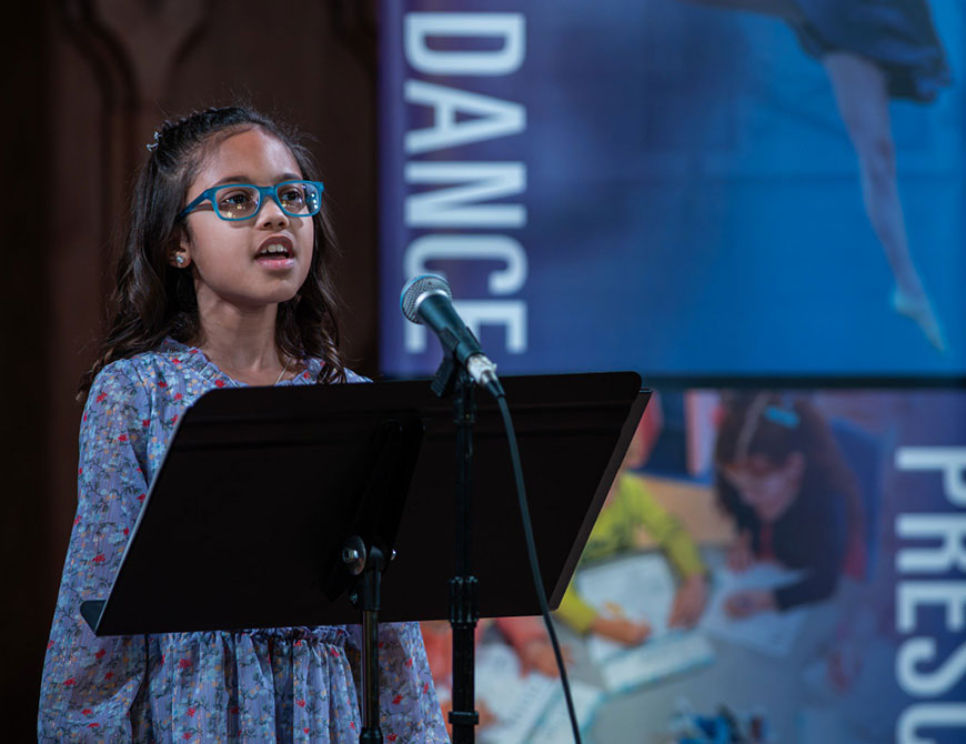 A child in a blue dress singing before a music stand and mic